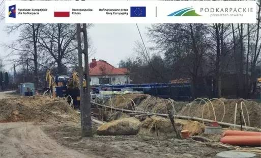 Grafika przedstawia prace budowlane związane z infrastrukturą podziemną, najprawdopodobniej z budową lub modernizacją sieci kanalizacji sanitarnej.  Na pierwszym planie widać wykopy ziemne, rury, przewody oraz elementy instalacyjne przygotowane do montażu