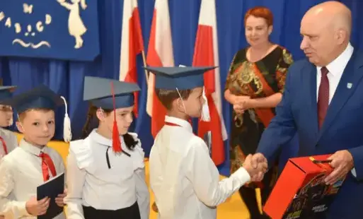 pasowanie na ucznia w szkole podstawowej w tarnogórze - burmistrza andrzej rychel wręcza uczniom teczki z przyborami szkolnymi w tle wychowawczyni anna sajak i czerwono-biało flagi